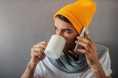 Bir fincan kahve, çay, su ile gri arka plan üzerinde oturan yakışıklı genç iş adamı portresi. Bir gadget'ı ile telefon konuşma. Turuncu şapka ve gri eşarp.