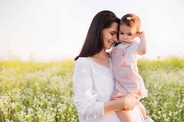Glück und Zärtlichkeit der Mutterschaft und Kindheit. hübsche brünette Mutter umarmt eine Tochter, Kleinkind, Baby in den Händen, lächelnd, auf Blumenfeld, Natur, Hintergrund. — Stockfoto