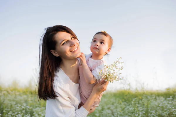 Красивый портрет матери обнимает дочь, малыша, ребенка, улыбаясь с цветами в руке, на цветочном поле, природе, небе фон. Материнство и детство . — стоковое фото