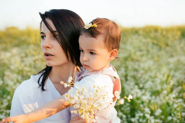 Pretty close up portrait of mother enbraces a daughter, toddler, baby, surprised and looking at one side with flowers in hand on field, nature background. Motherhood and childhood. — Stock Photo, Image