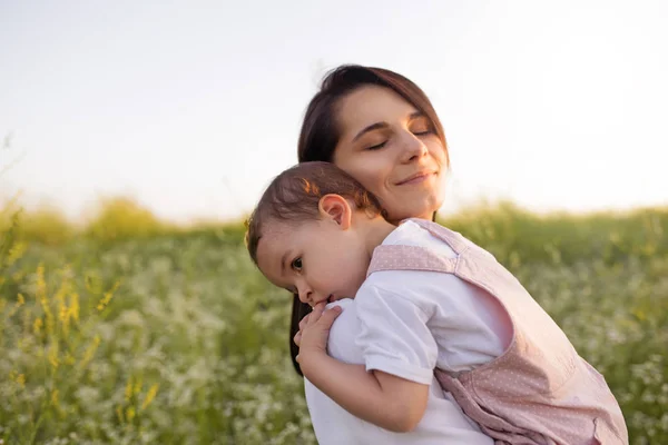 Schöne Mutterschaft, junge hübsche Mutter mit geschlossenen Augen umarmt eine Tochter, Kleinkind, Baby, auf den Händen, auf dem Feld, Naturhintergrund. — Stockfoto