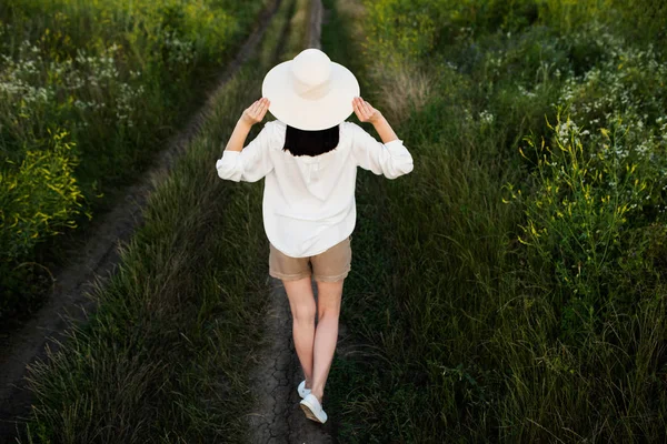 Um passado incrível. Linda e adorável, encantadora vista para trás da jovem mulher morena bonita com as mãos em um chapéu, andando sobre fundo campo verde . — Fotografia de Stock