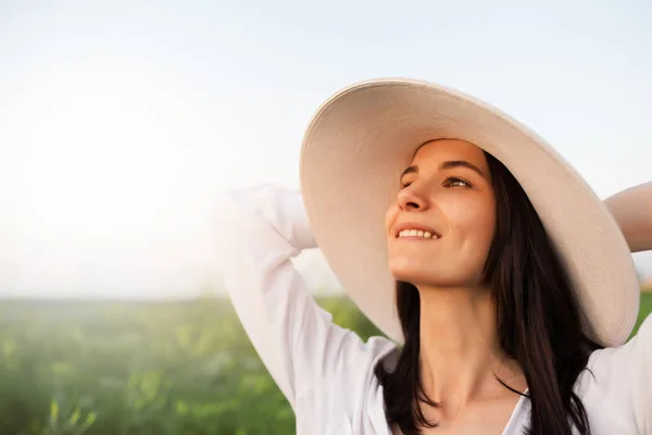 Close up belo retrato de uma jovem romântica e atraente com as mãos no chapéu e camisa branca, sonhando, olhando para o céu, na natureza, fundo de campo. Beleza — Fotografia de Stock