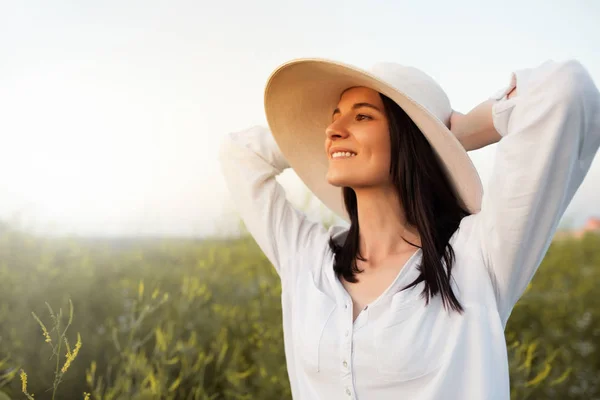 Красивый портрет романтичной и привлекательной молодой женщины с руками на шляпе и белой рубашке, мечтающей, смотрящей вперед на небо, на природу, на полевой фон. Красота — стоковое фото
