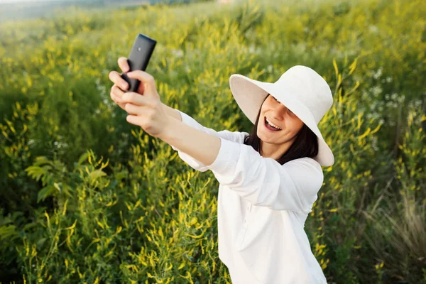 View from above of an young brunette beautiful smiling and laughing woman with white shirt taking selfie with a phone on nature, field background. Instagram. — Stock Photo, Image