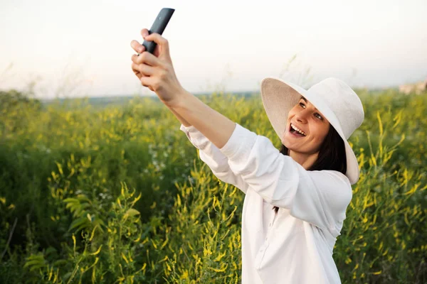 Vonzó gyönyörű romantikus fiatal nő, kalap és fehér ing mosolyogva, és figyelembe selfie a telefon, a természet, a háttér területen. Instagram — Stock Fotó