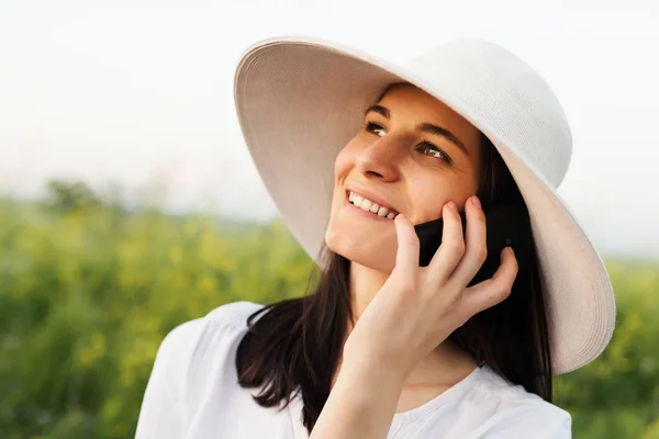 Portrait of beautiful attractive brunette young woman with a hat talking on the phone on nature, field background look up. — Stock Photo, Image