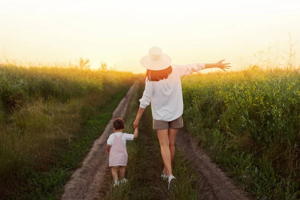 Материнство и детство. Удивительный, очаровательный, восхитительный вид симпатичной матери в шляпе с поднятой рукой и маленькой красивой дочки, идущей по дорожке поля. Закат или восход солнца . — стоковое фото