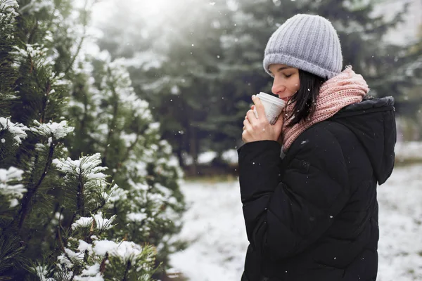 Güzel beyaz kadın kış ceket, bir fincan kahve ya da çay, açık havada park veya orman kış günü kar yağışı üzerinde tutarak gri bere şapka ve pembe eşarp, ördü. Kış ruh hali. — Stok fotoğraf