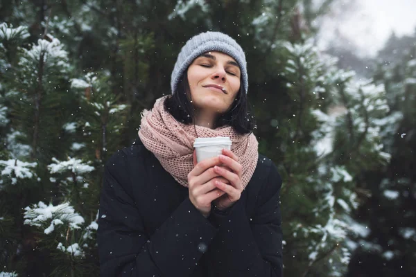 Güzel beyaz kadın kış ceket, bir fincan kahve ya da çay, açık havada park veya orman kış günü kar yağışı üzerinde tutarak gri bere şapka ve pembe eşarp, ördü. Kış ruh hali.