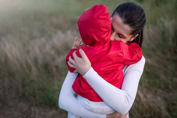Портрет красивой молодой матери, держащей своего малыша в красной куртке. Счастливая семья, мать и ребенок играют обниматься на поле прогулки на природе . — стоковое фото