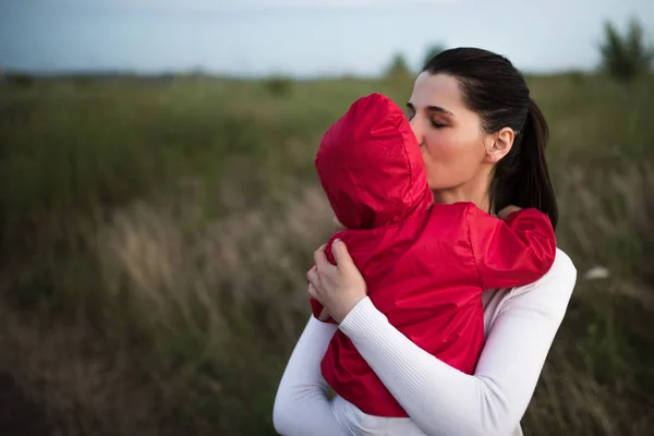 Bela jovem mãe segurando e beijando sua criança bebê usando jaqueta vermelha. Família feliz, mãe e criança brincam abraçando no campo caminham na natureza ao ar livre . — Fotografia de Stock