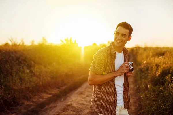 Bonito jovem viajante feliz com câmera vintage, em um fundo de prado. Humor de viagem. Fotografia. Relaxamento em um campo e por do sol. Explore a natureza . — Fotografia de Stock