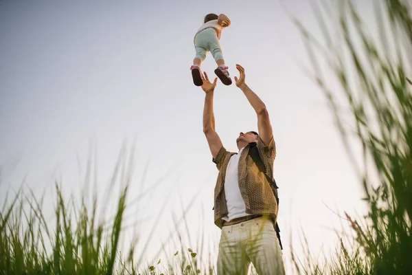 Przystojny młody ojciec rzuca swoją małą córeczkę w powietrze i łapie ją w ręce na świeżym powietrzu. Tata bawi się z dzieckiem w naturze. Szczęśliwa rodzina spędza razem czas na łące. — Zdjęcie stockowe