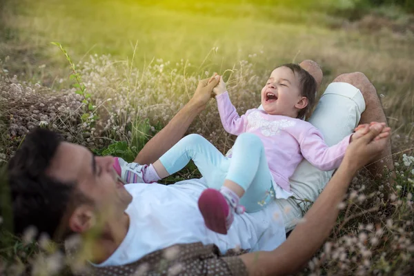 Piękny młody ojciec siedzi twarz do jego dziewczyna ładny maluch gry i śmiejąc się na trawie. Szczęśliwy ojciec z jego dziewczynka w słoneczny letni dzień. Rodziny pieszo na łące. — Zdjęcie stockowe