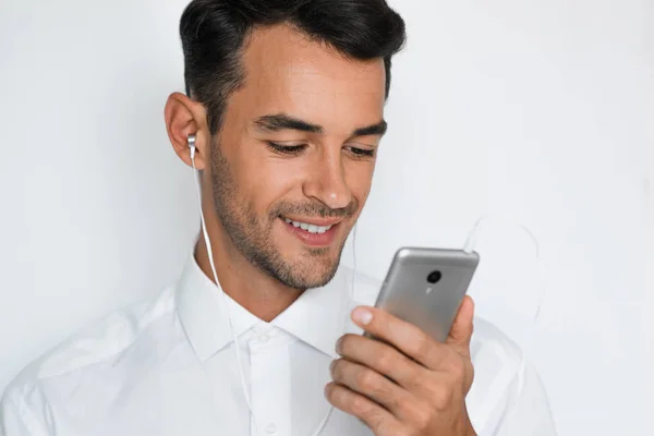 Retrato de un joven guapo y atractivo en camisa blanca con auriculares que sostienen el teléfono móvil aislado en el fondo gris claro. Feliz hombre de negocios con teléfono inteligente escuchar concepto de moda . — Foto de Stock