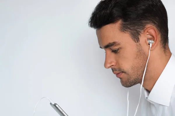 Tutup samping potret seorang pria muda tampan yang menarik dengan kemeja putih dengan earphone memegang ponsel terisolasi pada latar belakang abu-abu terang. Mendengarkan konsep bisnis — Stok Foto