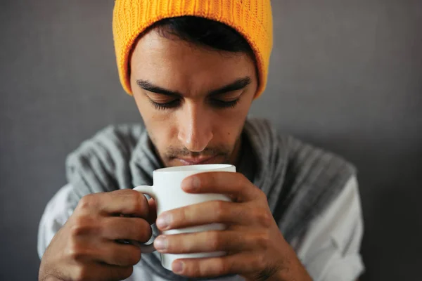 Close-up portrait of alone sitting young handsome man with a mug of coffee, tea, water, on gray background. Wearing orange hat and gray scarf. — Stock Photo, Image