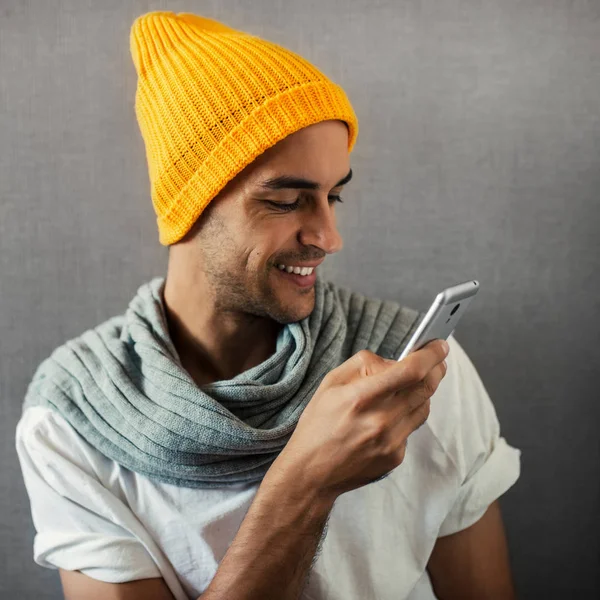 Улыбающийся красивый молодой человек в оранжевой шляпе и сером шарфе, с устройством, телефоном на сером фоне . — стоковое фото