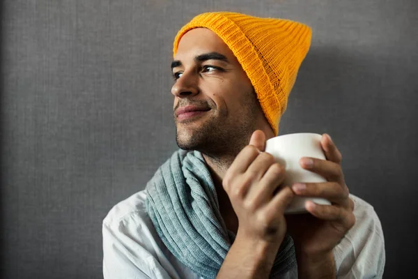 Dreaming, smiling and sitting young handsome man with a mug of coffee, tea, water, on gray background. Wearing orange hat and gray scarf. — Stock Photo, Image