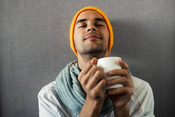 Sitting young handsome man with a mug of coffee, tea, water. Dreaming and smiling with closed eyes on gray background. Wearing orange hat and gray scarf. — Stock Photo, Image