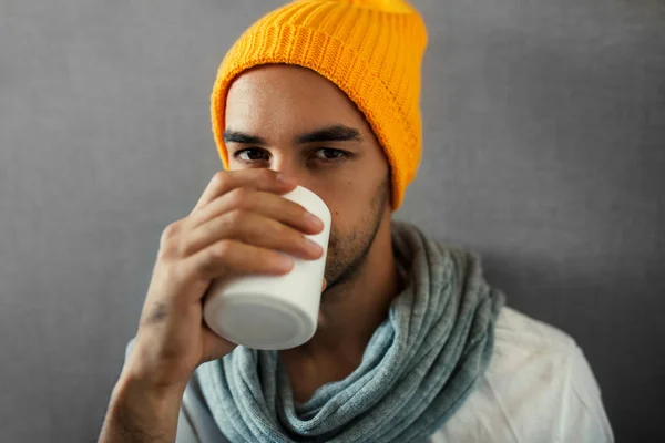 Handsome young man drinking coffee, tea, water, with white mug. Wearing orange hat and gray scarf in autumn or winter time, sitting on gray background. Looking at the camera.