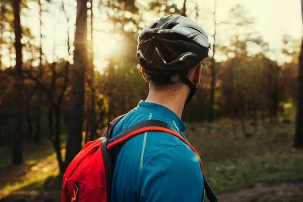 Close up shot from back of handsome young bearded man cyclist wearing protective helmet, blue t-shirt and red backpack, looking away, thinking about route and cycling. Travel concept. Sport lifestyle.