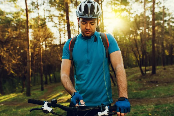 Young unshaven European cyclist in helmet, wearing blue cycling t-shirt, gloves switching speed mode on his white bike before riding uphill in forest. Travel lifestyle. Concentrated biking in mountain
