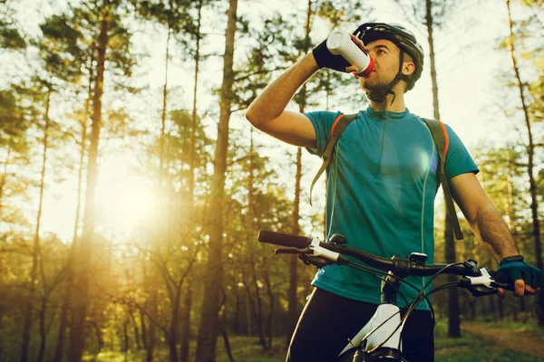 Όμορφος νεαρός επαγγελματίας ποδηλάτης ντυμένος με ρούχα ποδηλασίας και προστατευτικό κράνος αισθάνεται ελεύθερος και χαρούμενος, πόσιμο νερό από το μπουκάλι, απολαμβάνοντας ποδήλατο στη ζεστή άνοιξη. Αναψυχή του αθλητή. — Φωτογραφία Αρχείου