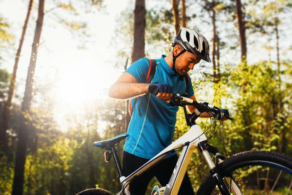 Handsome serious and concentrated young european cyclist wearing blue cycling t-shirt, helmet, gloves and red backpack switching speed mode on his white bike before riding uphill. Travel lifestyle. — Stock Photo, Image