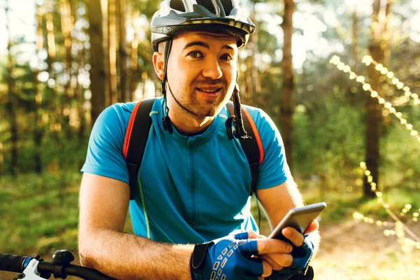 Retrato del joven ciclista caucásico parado en el bosque, sosteniendo el teléfono móvil, utilizando la aplicación en línea para buscar coordenadas GPS mientras monta bicicleta. Viajes, deporte y tecnología moderna . —  Fotos de Stock