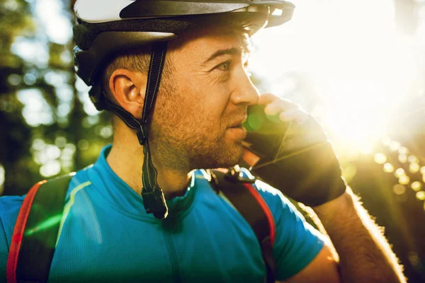 Close up portrait of young Caucasian cyclist in sportswear wearing helmet and blue t-shirt, talking on mobile phone with friend looking away, having small break while exercising outdoors in forest. — Stock Photo, Image