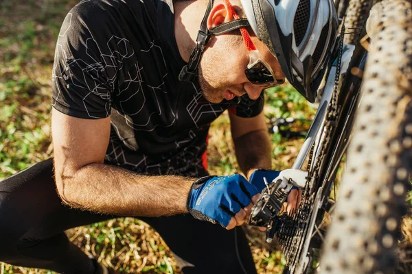 Attractive outdoor close up shot of concentrated young rider in protective gear sitting on grass in front of his broken bicycle, trying to figure out what's the problem. — Stock Photo, Image