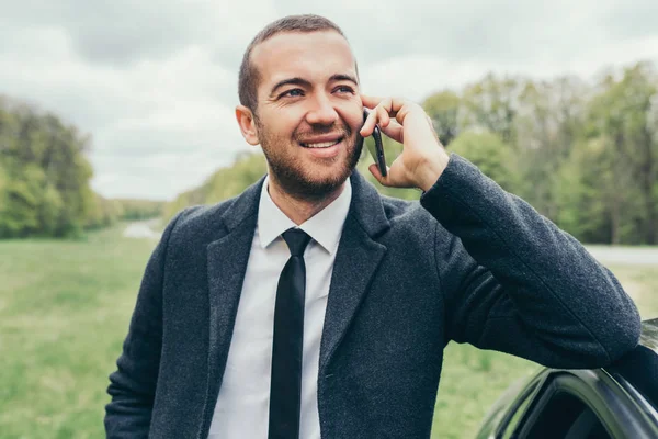 Çekici Avrupa beyaz Yöneticisi'nde meslektaşları test sürüşü için beklerken arabasını yakın cep telefonuyla şık resmi ceket. Şık mutlu gülümseyen iş adamı var akıllı telefon konuşma.