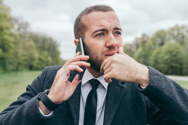 Portrait of handsome young urban European professional man using mobile gadget outdoors. Trendy looking entrepreneur making business call, talking on smart phone to his partner, having serious look.