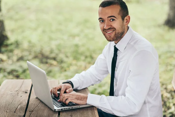 Lachende knappe man in een wit overhemd zit op houten tafel met behulp van laptop hebben verheugd oprechte glimlach tijdens het surfen op Internet. Mensen, carrière, emoties, technologie concept. — Stockfoto