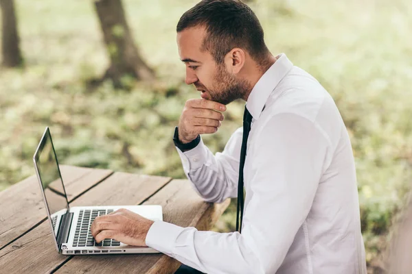 Perfil de toma de serio y concentrado joven hombre de negocios caucásico sentirse frustrado, sentado al aire libre con PC portátil genérico, tocando la barba, tratando de concentrarse en el trabajo, buscando agotado . — Foto de Stock