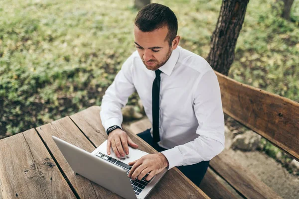 Pohled shora chytrý podnikatel nosit bílou košili, psaní na počítači dělat zpráva jeho firmy. Pohledný muž pracuje na notebooku venkovní v parku. Koncept technologie, okupace a práce. — Stock fotografie