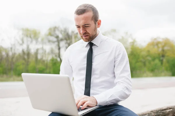 Moda homem de negócios inteligente vestindo camisa branca, digitando em seu computador fazendo relatório de sua empresa usando wireless. Bonito homem trabalhando em laptop ao ar livre no parque. Conceito de tecnologia e negócios . — Fotografia de Stock