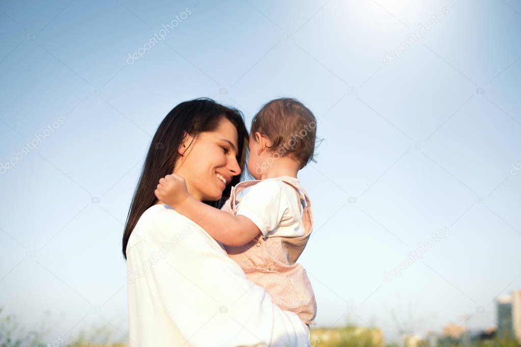 Beautiful happy motherhood, young pretty mother smiling, looking and embraces a daughter, toddler, baby, on hands, on field, sky, background.