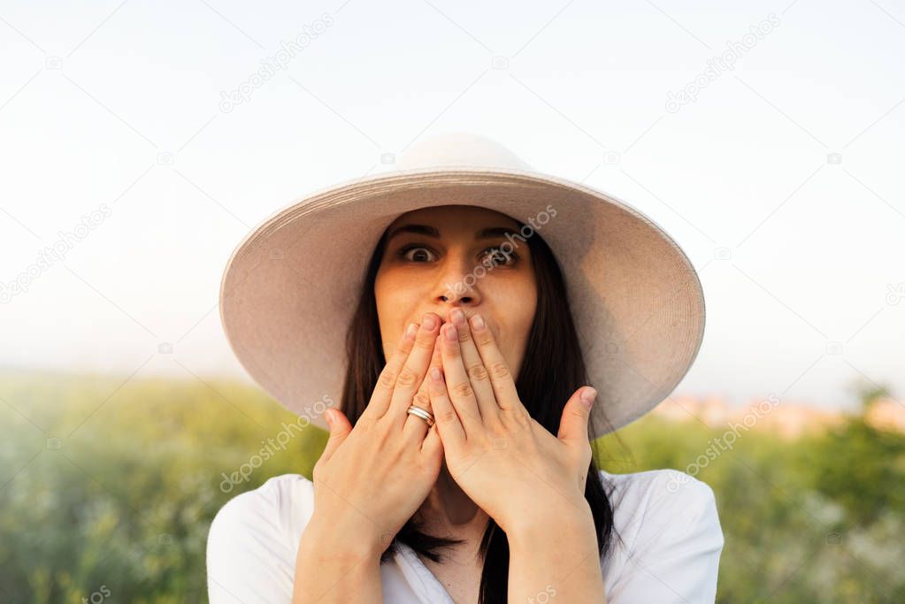 Beautiful portrait of an attractive brunette young woman with a hat surprised or shocked with hands on the mouth, on nature, field background look up.