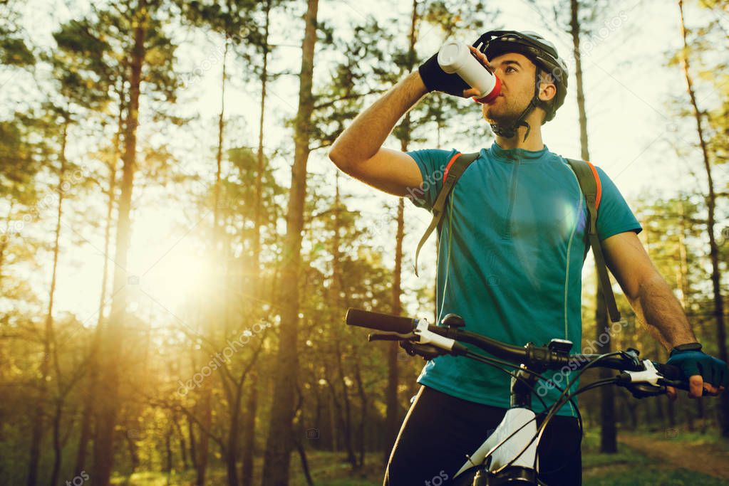 Handsome young professional cyclist dressed in cycling clothing and protective helmet feeling free and happy, drinking water from the bottle, enjoying bicycle in warm spring. Recreation of sportsman.