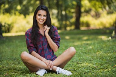 Çekici gülümseyen ve park doğa çerçevede ekose gömlek siting giymiş mutlu genç esmer beyaz kadın doz vücut. Yaşam tarzı, insan ve güzellik kavramı.