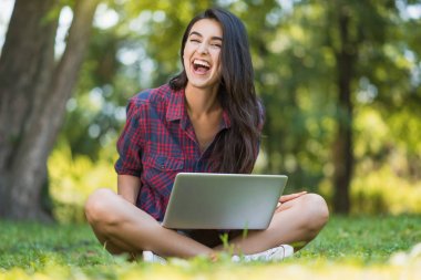 Genç Beyaz mutlu kadın gülüyor ve çim dizüstü pc, önünde oturan giyen ekose gömlek çekici atış dizüstü bilgisayarında, alışveriş ve kablosuz iletişim çalışma.