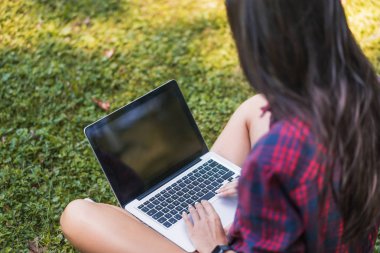 Alışveriş ve eğitim ekose gömlek giymiş, çim dizüstü pc önünde oturan, kablosuz iletişim ile onun dizüstü bilgisayar üzerinde çalışan genç beyaz esmer bir kadın üst arka görünümü.