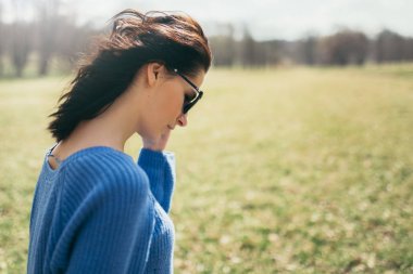 Güzel genç esmer kadının yan görünüm portre saç doğa arka plan üzerinde esen rüzgar dönüyoruz. Çekici kız parkta güneş gözlüklü. Yaşam tarzı kavramı. Kapak fikir konsepti.