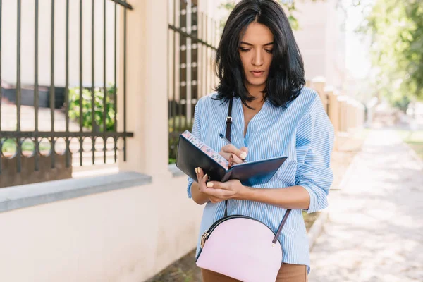 Tiro horizontal de mulher de negócios séria gerencia finanças, mantém o bloco de notas em mãos, olha atentamente no caderno, andando pela rua na cidade. Estilo de vida, pessoas e conceito de tecnologia — Fotografia de Stock