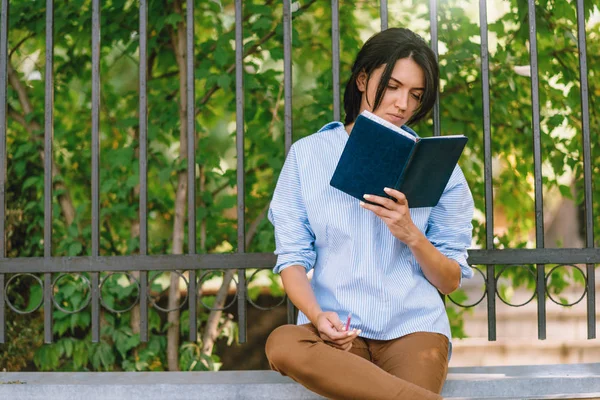 Серйозна жіноча модель зосереджена на читанні, сидить на стіні в парку. Захоплений студент готується до майбутньої сесії в коледжі. Люди і концепція способу життя . — стокове фото