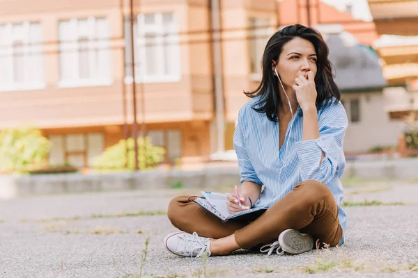 Brunette European beautiful woman sitting at sidewalk, studying and making notice on notebook planner. Young cute woman with earphones listen music and preparing for exam. People and lifestyle concept