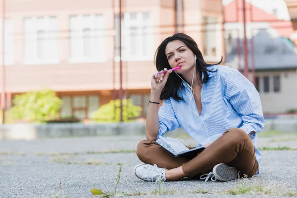 Mulher bonita caucasiana sentado na calçada, estudando e fazendo notar no planejador notebook. Jovem com fone de ouvido ouvir e se preparando para o exame . — Fotografia de Stock